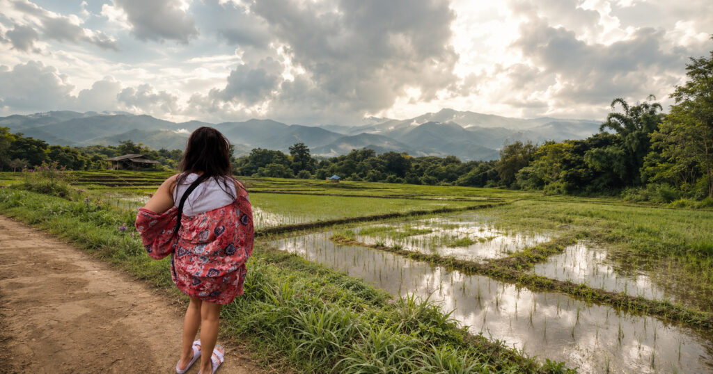 Woman in a coral kimono looking out over flooded rice paddies toward the misty Mae Hong Son mountains in Pai, Northern Thailand